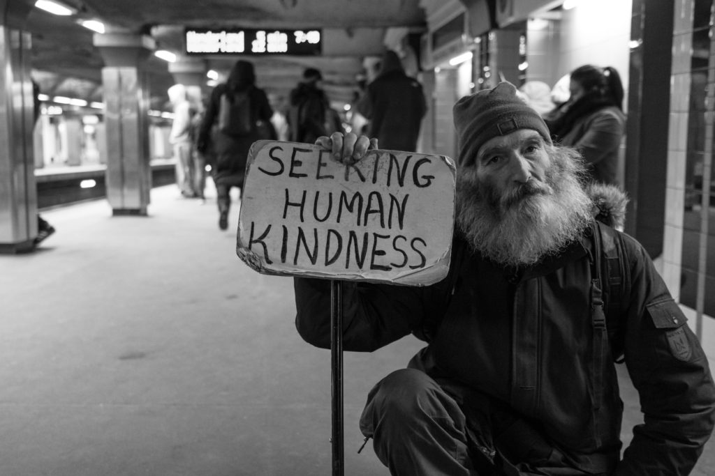 Homeless man holding a sign that reads Seeking Human Kindness